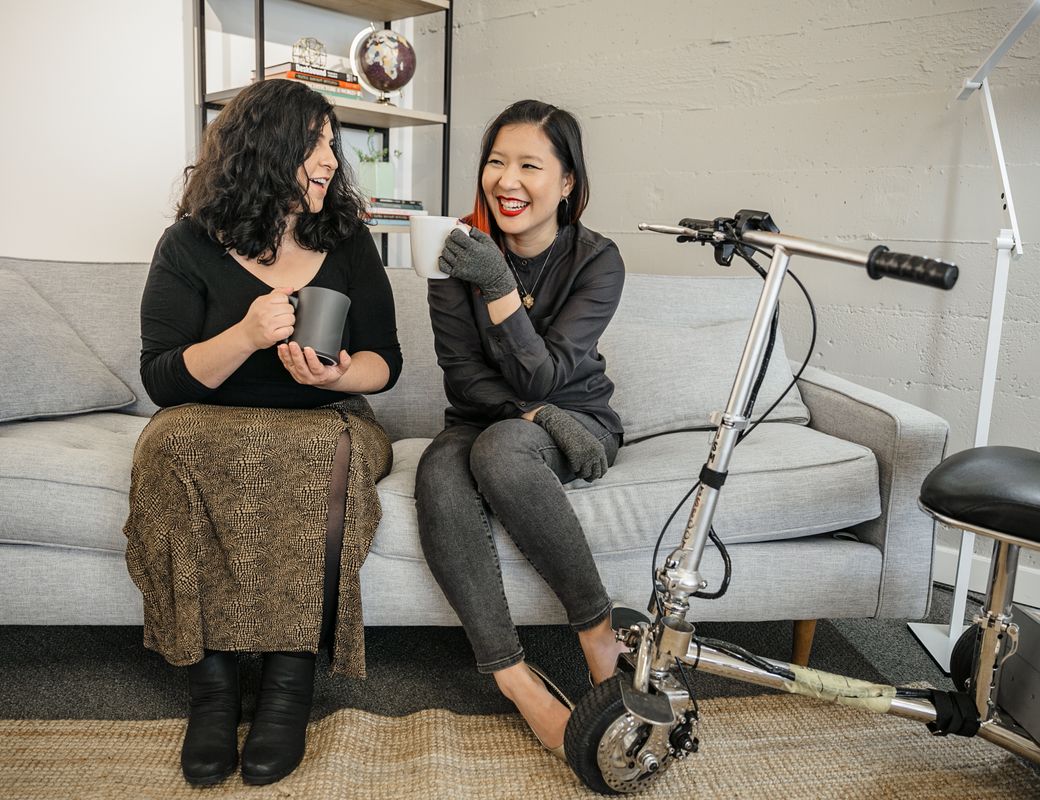 A Latinx disabled woman and an Asian disabled genderfluid person chat on a couch, both holding coffee mugs. A lightweight mobility scooter rests to one side.