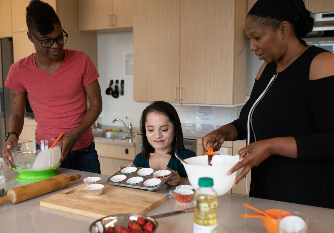 Three disabled people of colour baking together in a kitchen — one holds a mixing bowl, one prepares a muffin pan, and one measures out a chocolate mix.