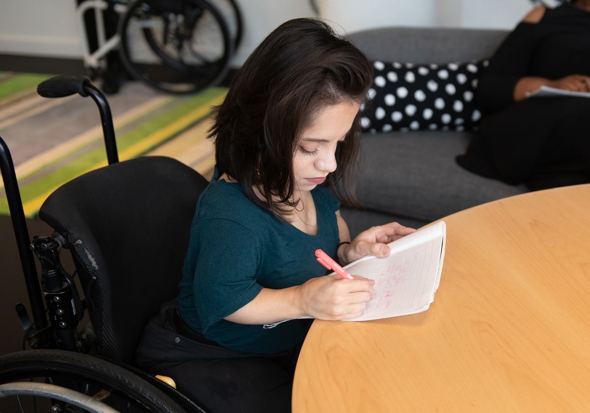 A South Asian person in a wheelchair takes notes by hand during a meeting.